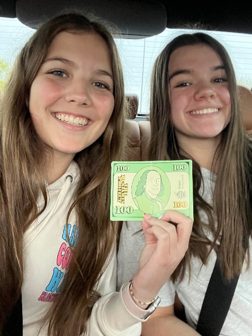 Brooklyn and Paisley holding a Benjamin Project $100 card, smiling in a car