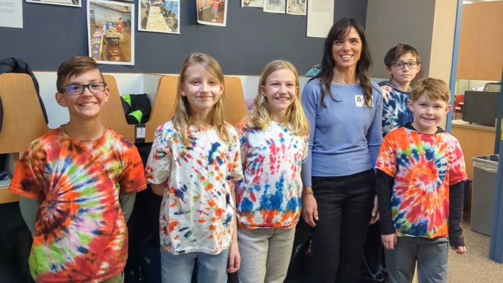 Sarah, a Global Doing Good Education Ambassador, stands with a group of students in Tie-Dye shirts in front of Roxbourgh Intermediate’s  “Benjamin Wall”