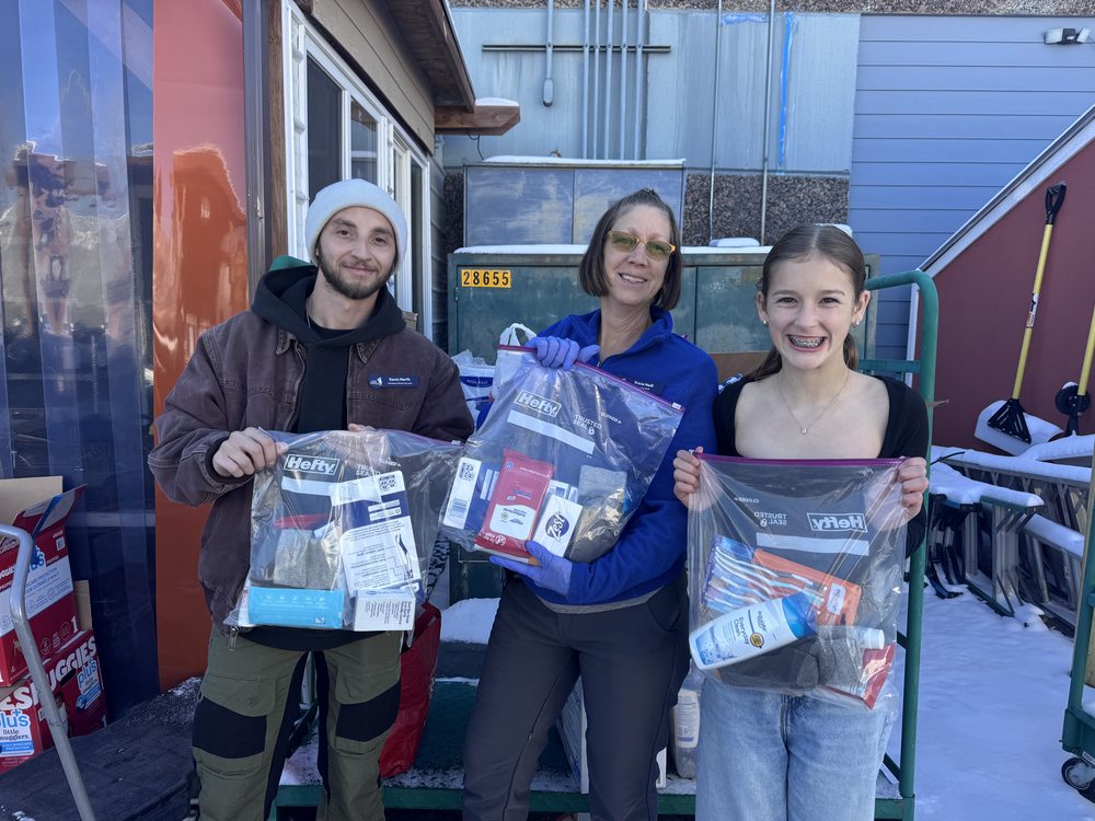 Two students and an adult volunteer standing outdoors, each holding up a clear zip-top bag of hygiene supplies