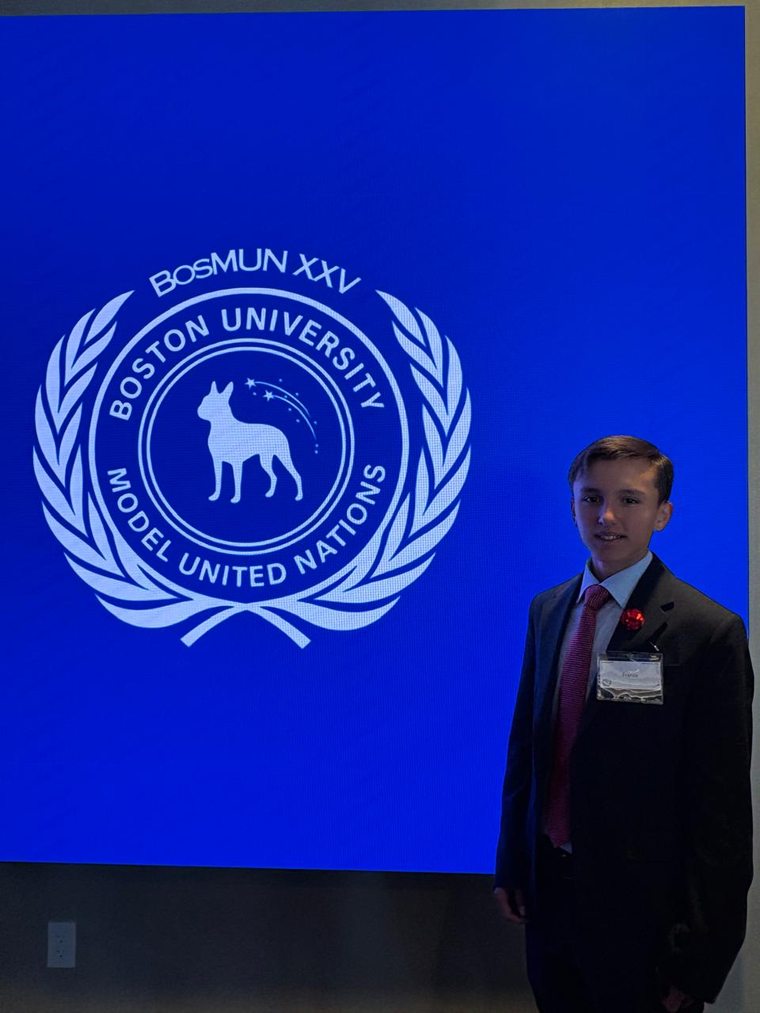 A student in a suit standing beside a large screen displaying the Boston University Model United Nations (BosMUN XXV) emblem