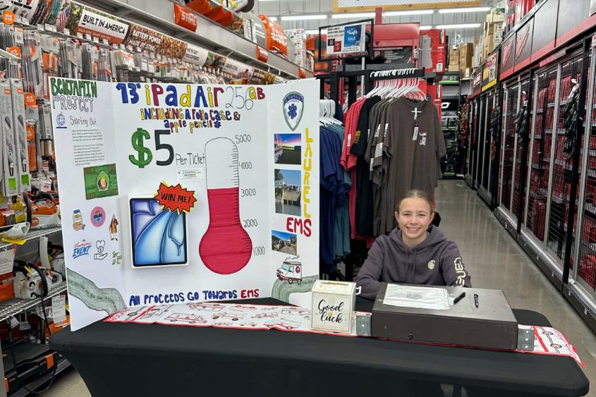 Claire at her raffle booth inside the local hardware store, with a hand-drawn poster advertising the iPad Air raffle for her local EMS