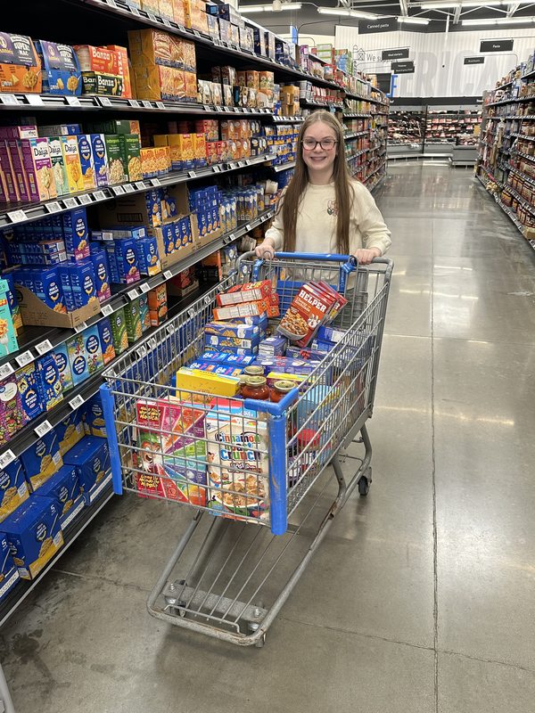 McKenzie pushing a cart full of food and supplies through the store aisle
