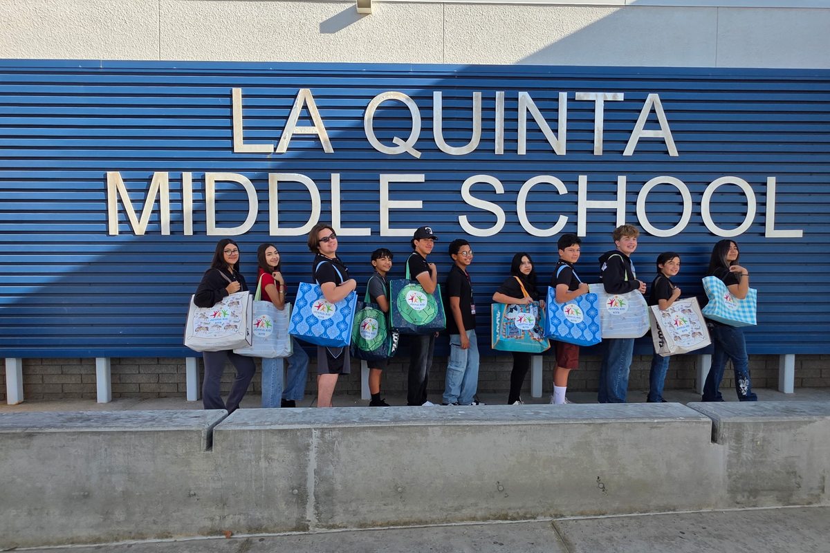 A group of middle school students holding decorated comfort bags in front of their school sign