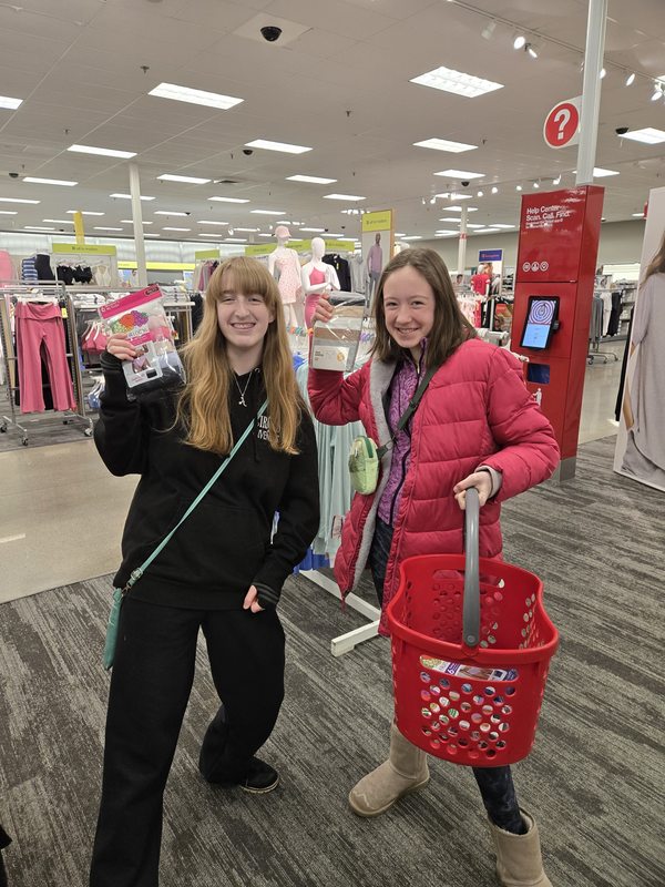 Two students shopping for supplies at a store, one holding a package and the other carrying a red shopping basket