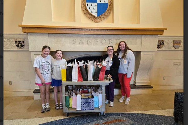 Two students and two hospital staff members with a cart of gift bags in front of the engraved Sanford fireplace