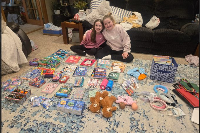 Two students seated on the floor behind a large spread of donated toys, games, coloring books, and stuffed animals