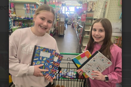 Two students smiling at a store, holding notebooks and coloring books over a cart full of supplies