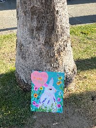 A brightly colored painting of a figure surrounded by flowers, propped against a tree trunk in a public space
